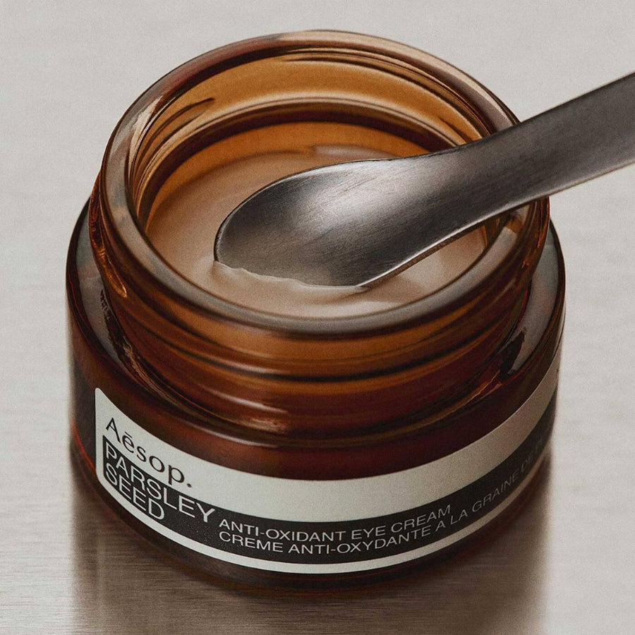 An open amber glass jar of Aesop Parsley Seed Anti-Oxidant Eye Cream with a metal spoon resting inside, set against a neutral light gray background.